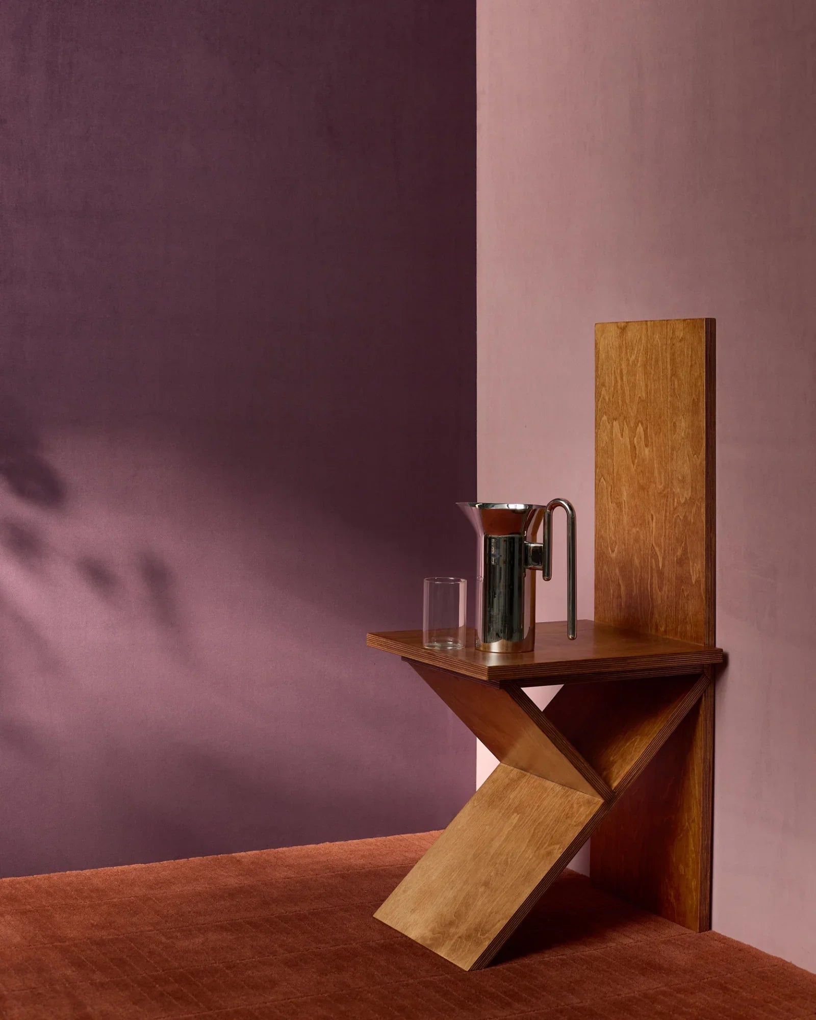 Modern geometric wooden side table with a glass and metallic pitcher against purple and pink walls