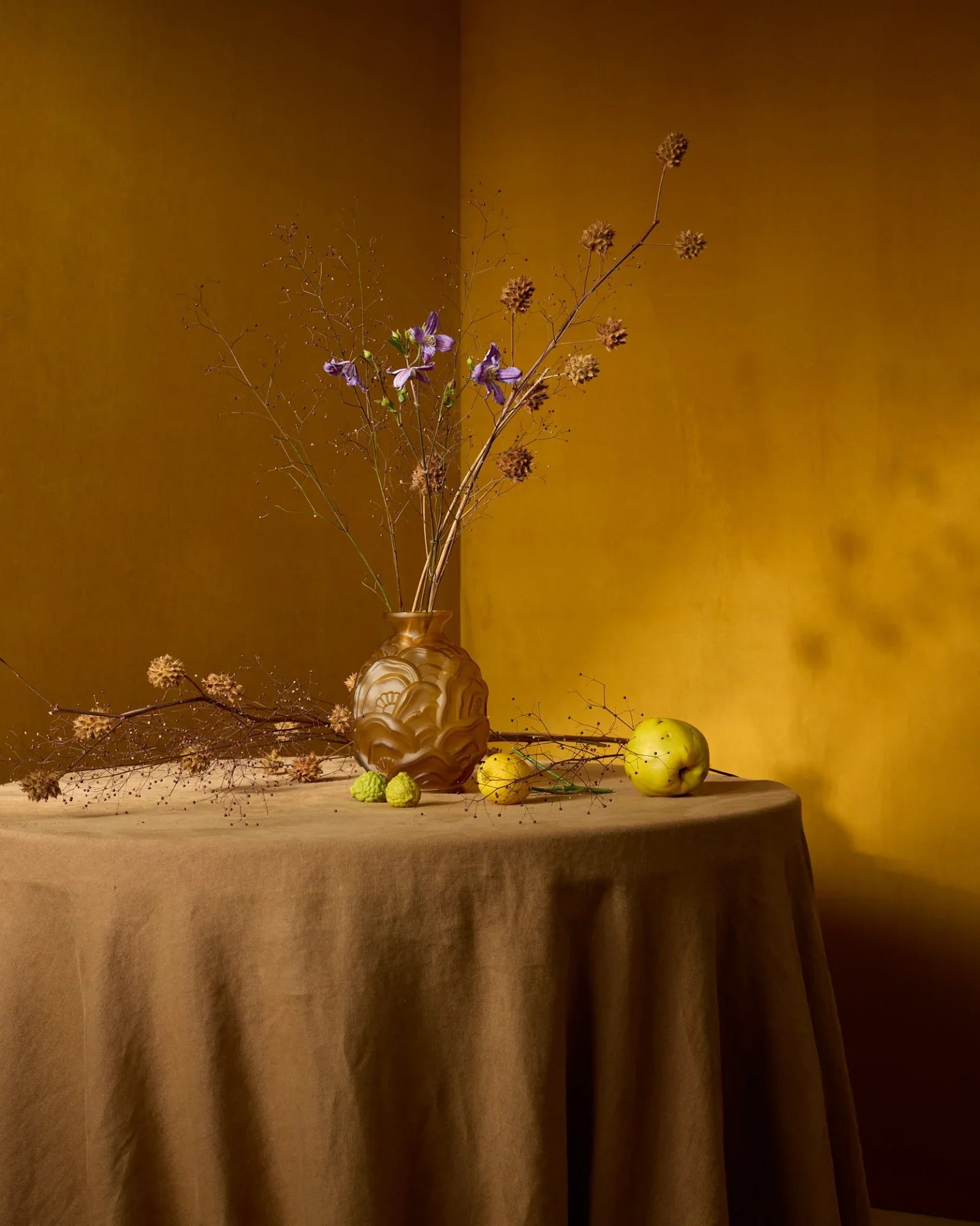 Elegant still life with yellow ceramic vase, dried branches, and fruit on a brown tablecloth.