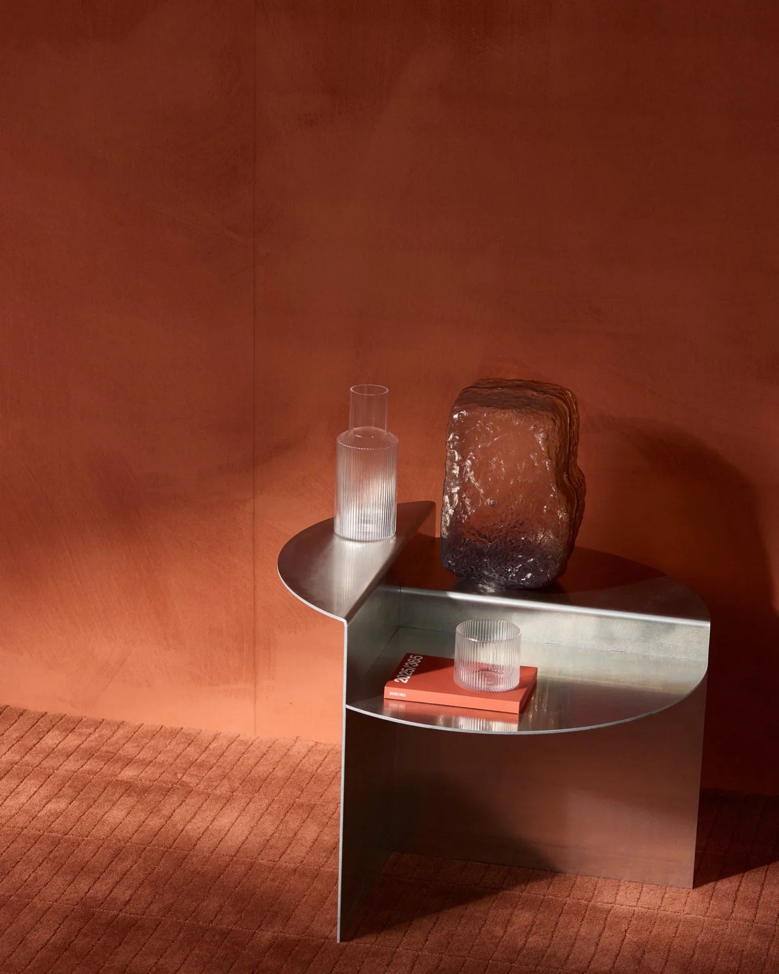 Modern silver metal side table with glass vase, two ribbed glasses, and a book in a minimalist interior with terracotta walls and carpet.