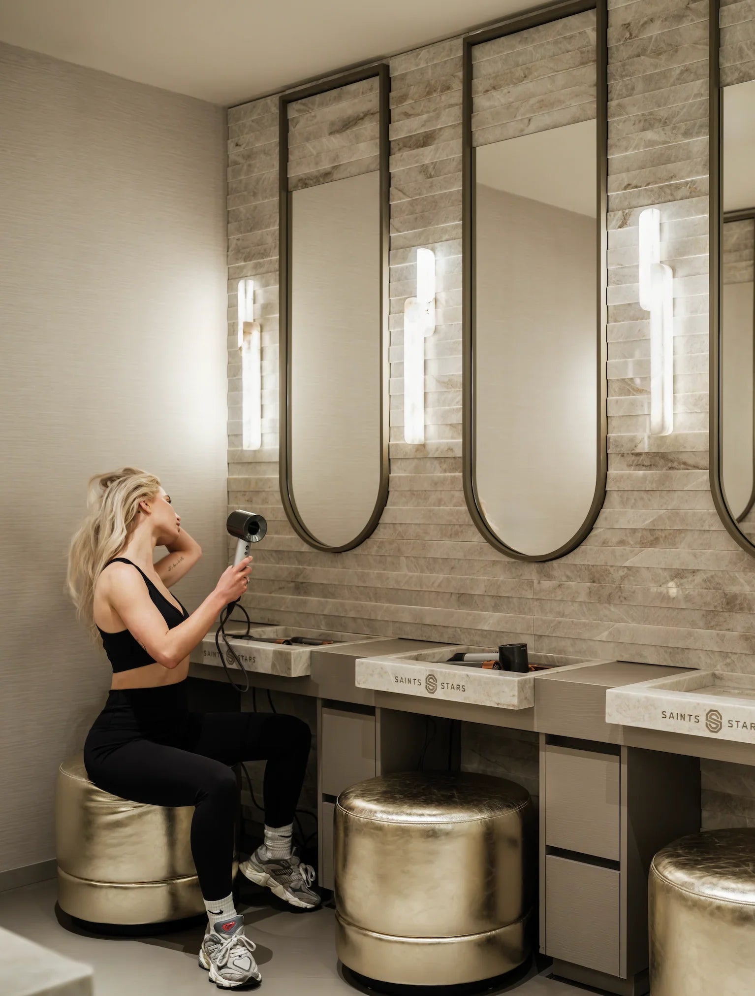 Woman blow-drying hair in modern luxury vanity area with marble accents and gold stools