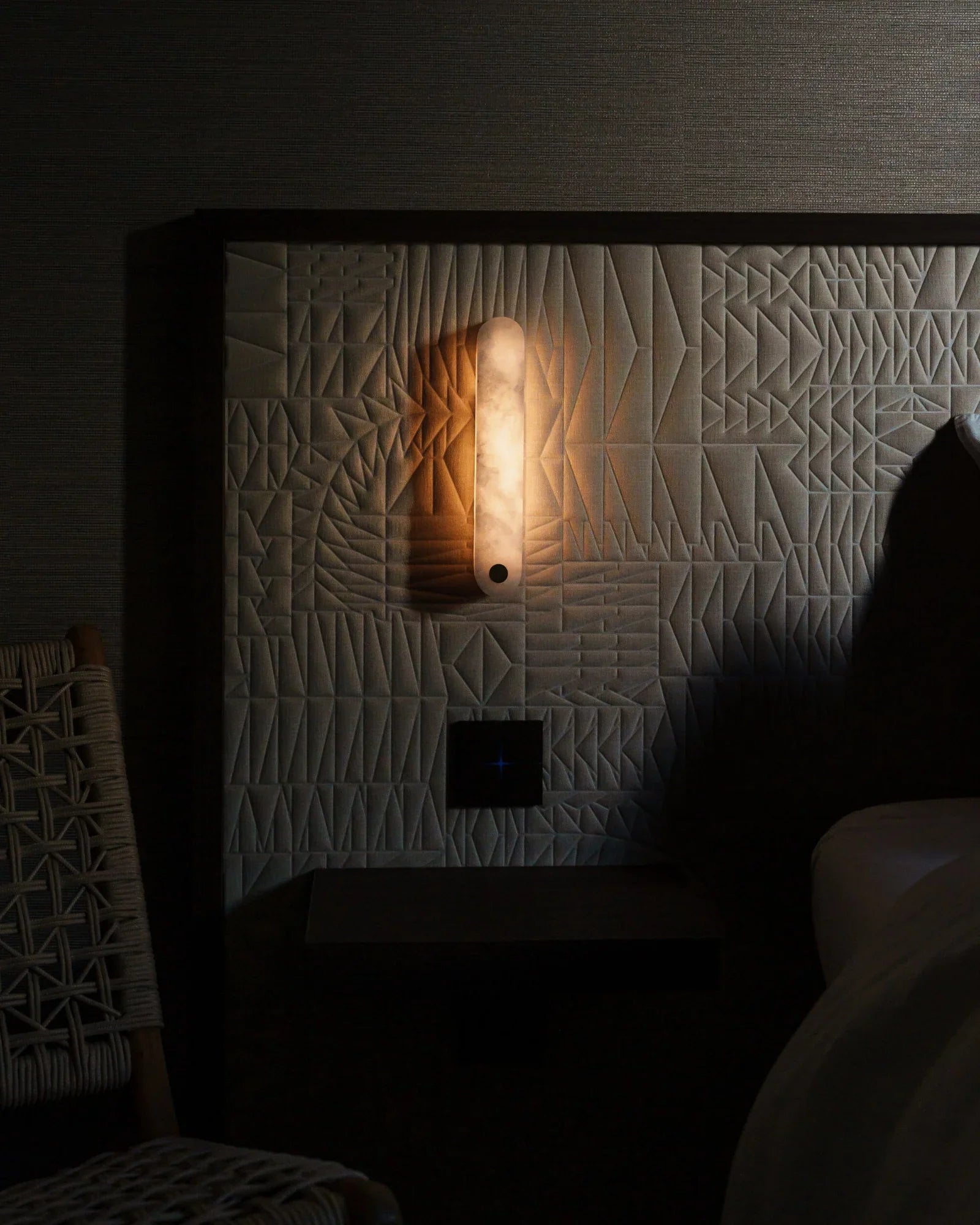 Modern bedroom with textured wall panel, glowing wall sconce, and woven chair