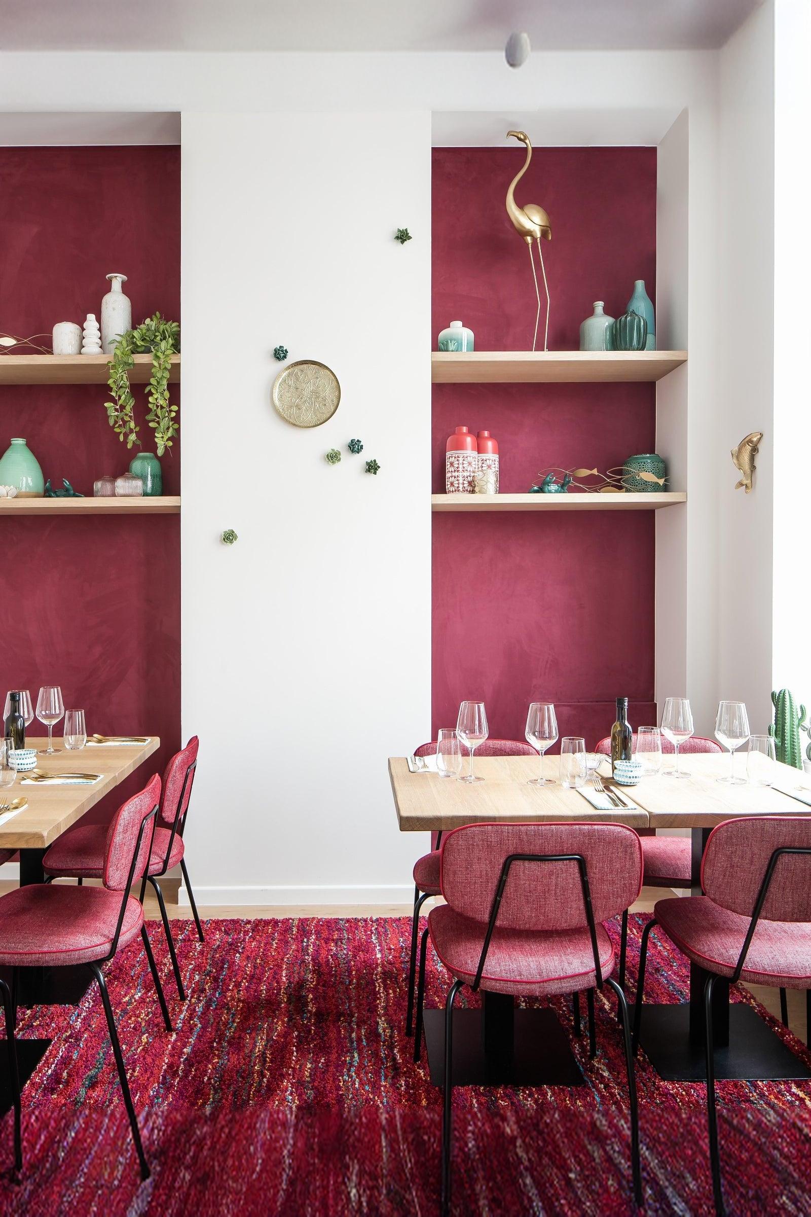 Modern dining area with pink chairs, wooden tables, red accent wall, and decorative shelving.