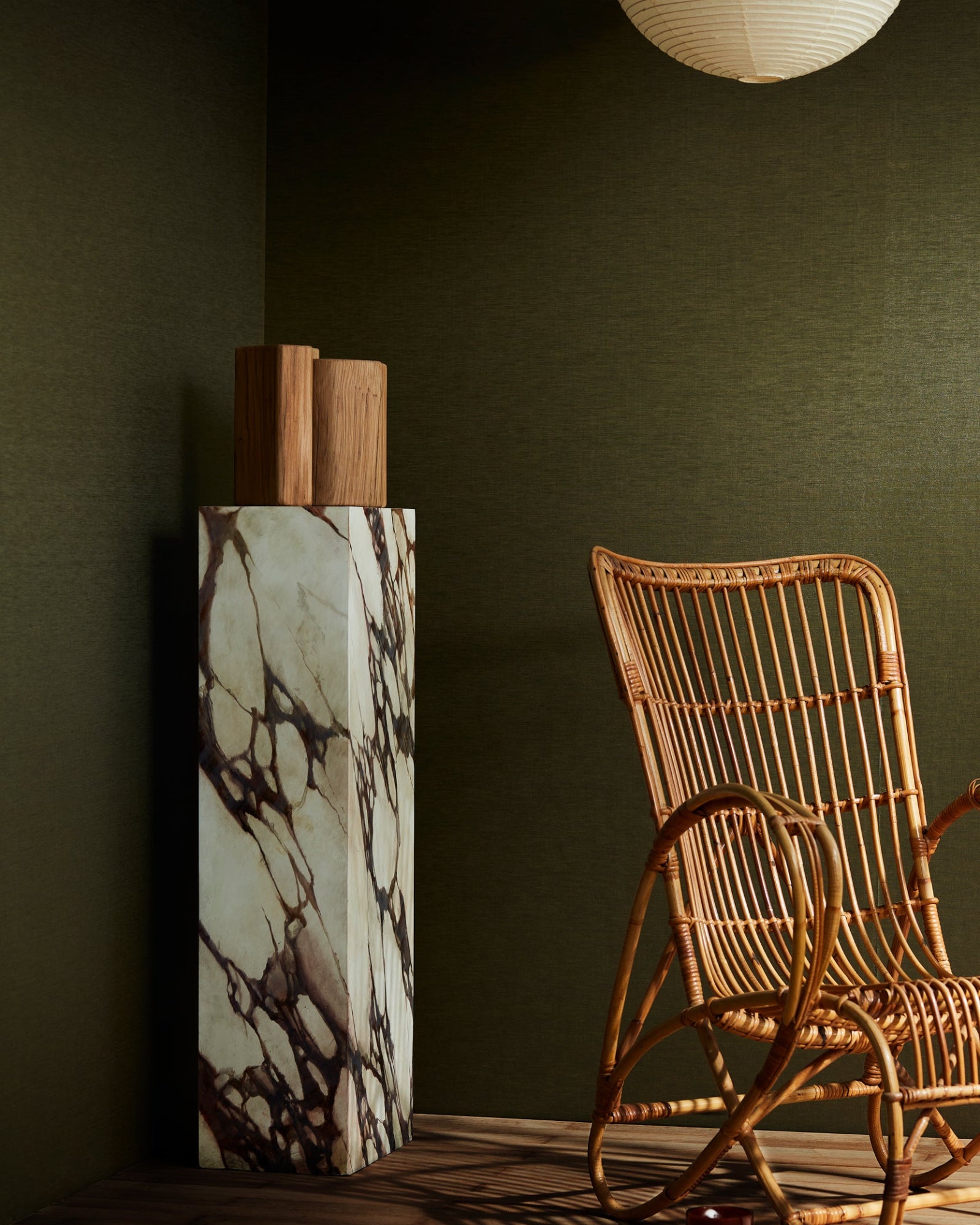 Luxury interior with rattan chair, marble pedestal, wood decor, and olive green wall