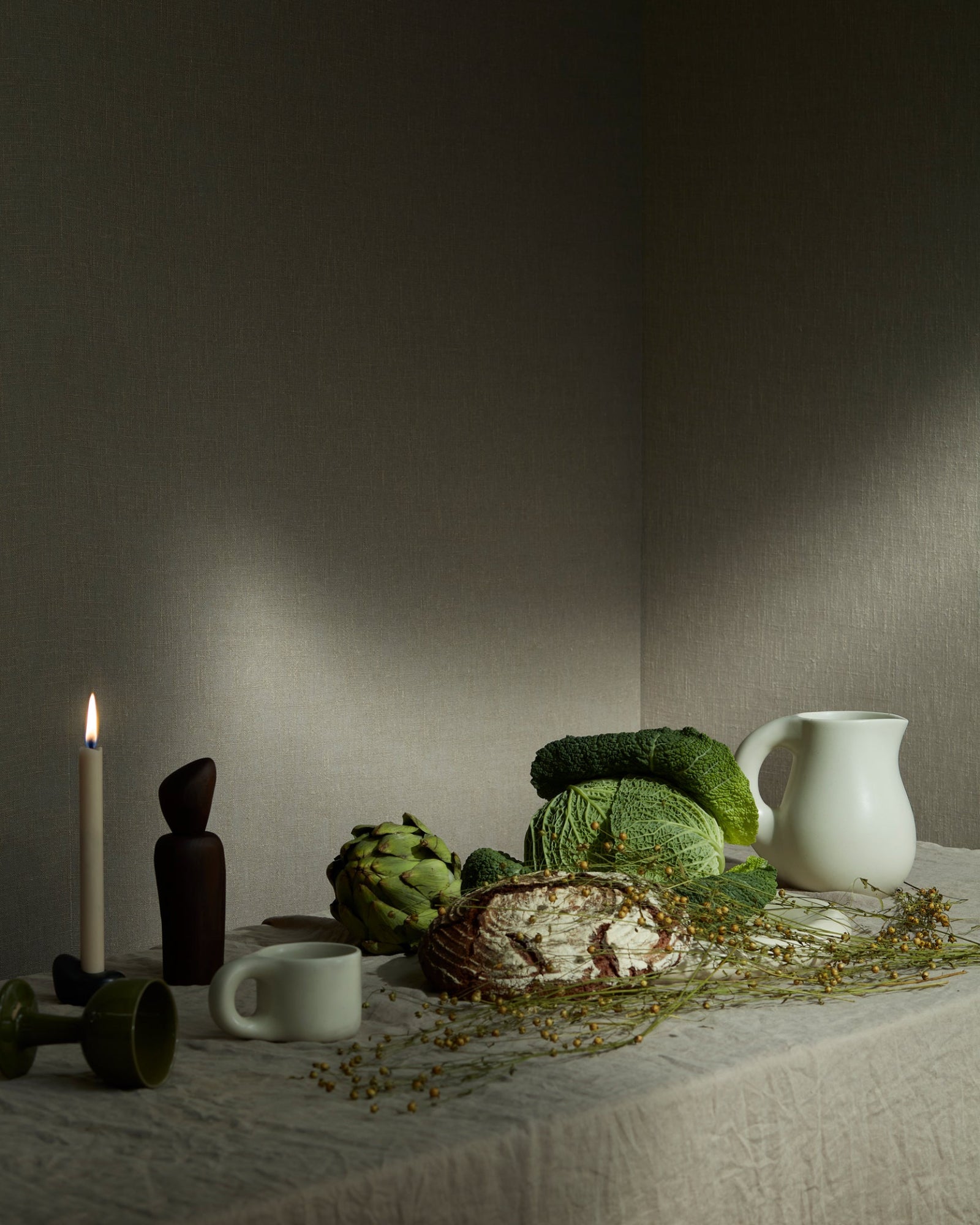 Elegant still life with artisanal bread, cabbage, artichoke, candle, and ceramic pitcher on a linen tablecloth