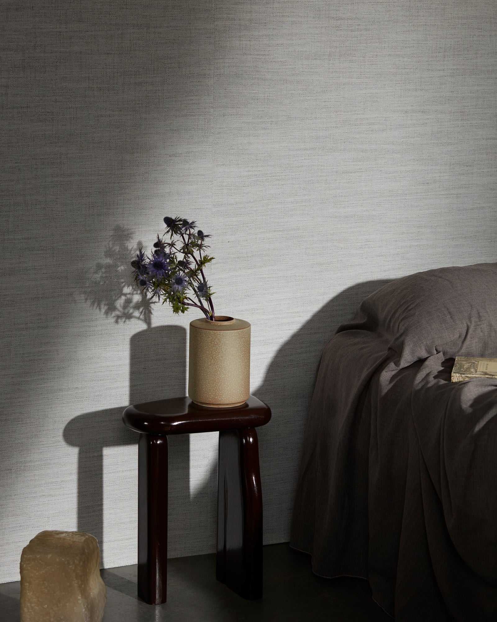 Minimalist bedroom with textured wall, dark wood side table, ceramic vase, and purple flowers.