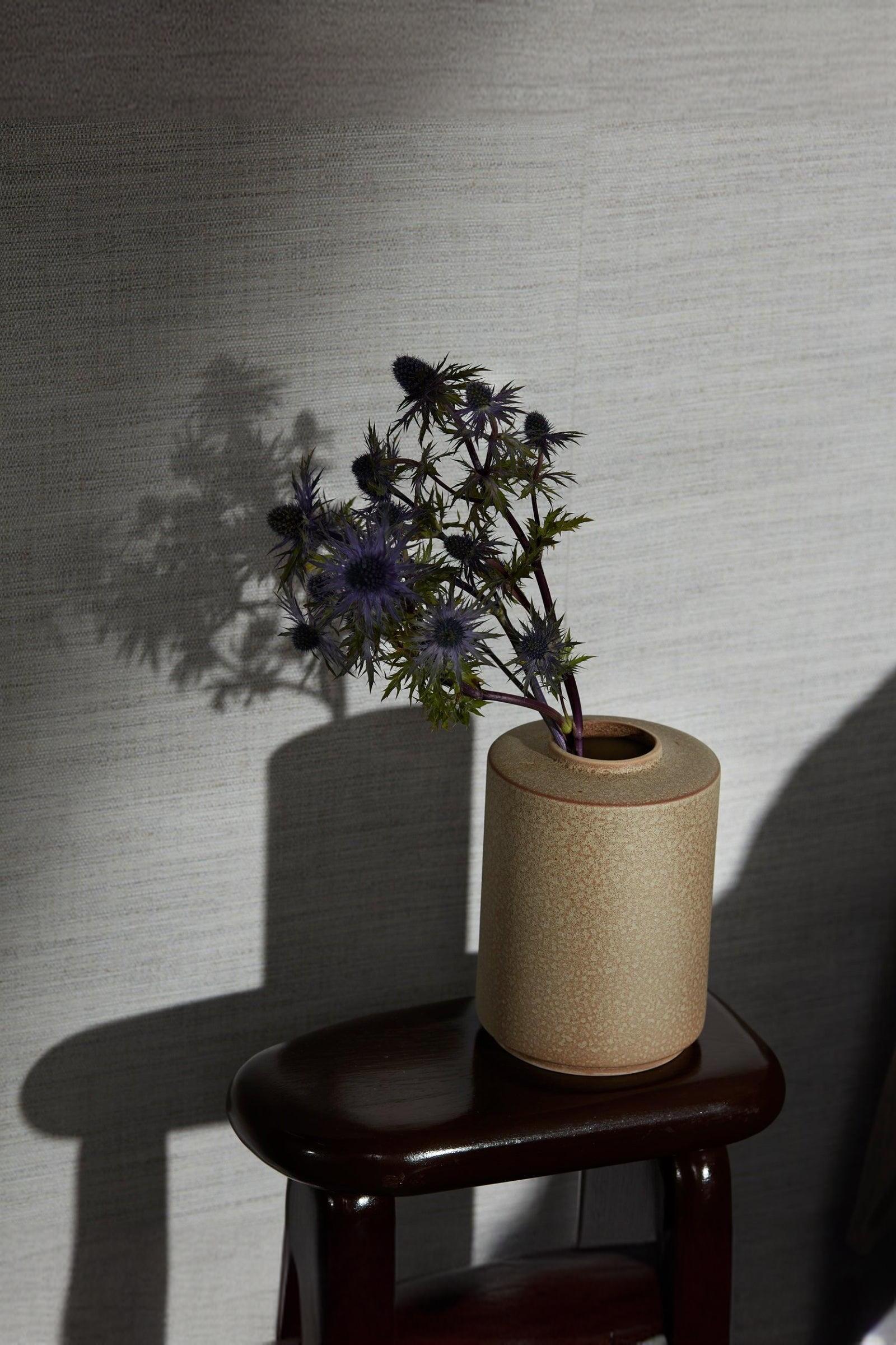 Beige ceramic vase with purple thistle flowers on a dark wood stool, minimalist interior
