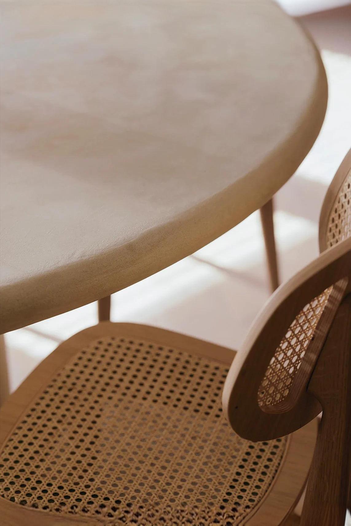 Close-up of a round light wood table and a woven rattan dining chair in a sunlit room