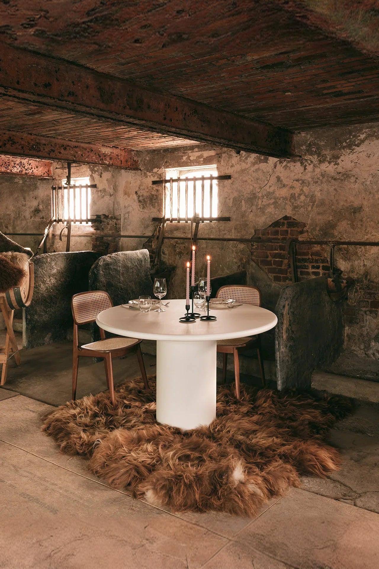 Modern round dining table with cane chairs, candles, and fur rug in a rustic stone interior